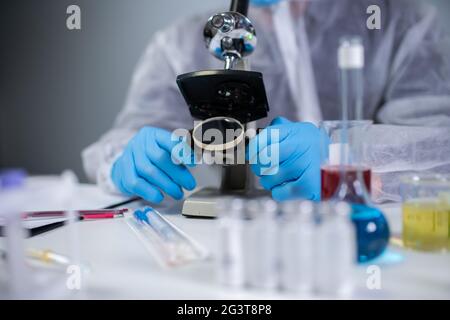 Medizinischer Fachmann in weißem Schutzanzug, der im chemischen Labor durch das Mikroskop schaut. Suchen Sie nach Impfstoff-Coronavirus. Cov Stockfoto