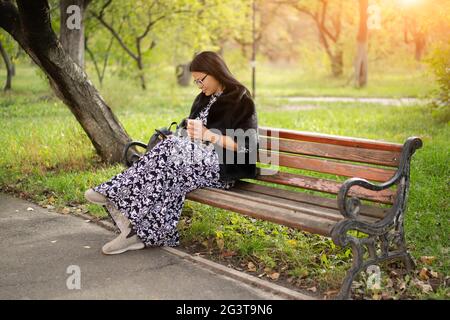Frau asiatischer Herkunft in schönem langen Kleid sitzt im Park auf einer alten rostigen Bank und liest Buch mit ihrem Gadget oder überprüft Socia Stockfoto
