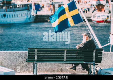 Stockholm, Schweden. Old man sitzt auf EINER Bank in der Nähe der Stadtpromenade. Im Hintergrund EINE schwenkende schwedische Flagge. Stockfoto
