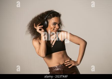Fröhliches, junges afroamerikanisches Mädchen, das Musik hört, trägt ein schwarzes Oberteil und eine Lederhose in Kopfhörern, isoliert auf grauem Rückenband Stockfoto