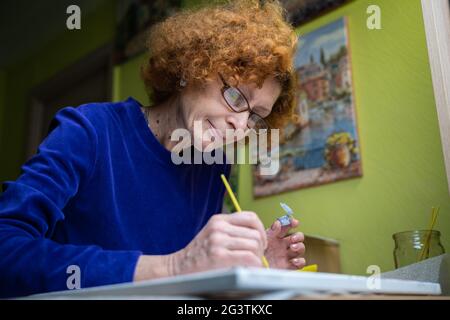 Glücklich reifen Künstler Frau Malerei auf Leinwand zu Hause Studio. Kreativität zum Ausdruck bringen. Positive ältere Frau mit Pinsel und drawin Stockfoto