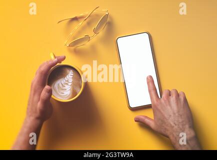 Der Mann, der ein Smartphone anheucht, verspottet auf einem hellgelben Schreibtisch mit Kaffeetasse und Gläsern Stockfoto