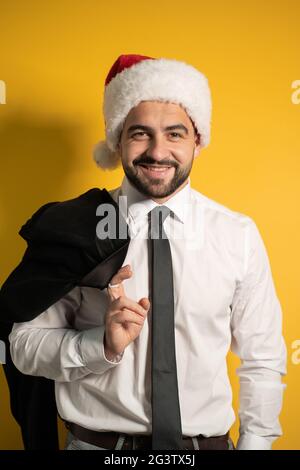 Bärtig lächelnder, hübscher Geschäftsmann im Weihnachtsmann-Hut, der eine graue Suite trägt und seine Jacke auf der Schulter hält, um sie zu hängen Stockfoto