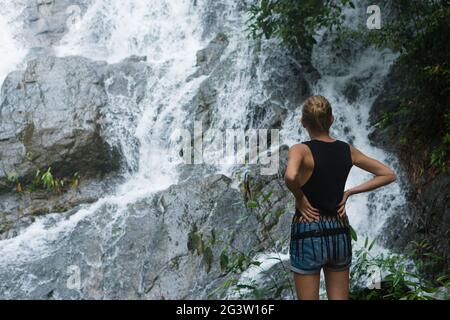 Blonde Frau, die auf einem Felsen steht und in der Nähe des Wasserfalls aufschaut Stockfoto