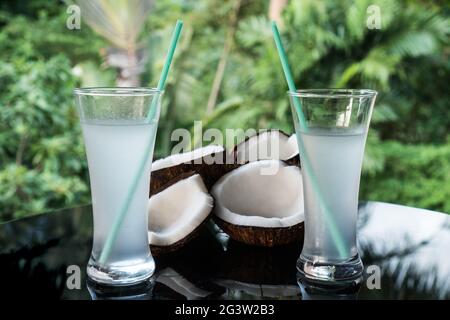 Kokosnüsse und Kokoswasser auf dem schwarzen Glastisch, isoliert über verschwommenem Palmenhintergrund Stockfoto