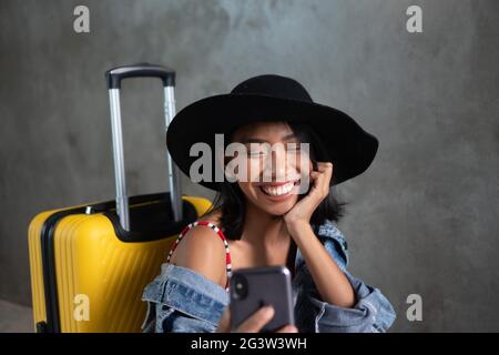 Porträt einer lächelnden jungen asiatischen Frau mit Gepäck und Mobiltelefon isoliert über Betonwand Stockfoto