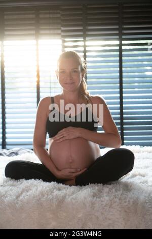 Glückliche, schwanger Frau in schwarzen Leggins und BH, die ihren Bauch hält, während sie sich zu Hause auf ihrem Bett entspannt Stockfoto
