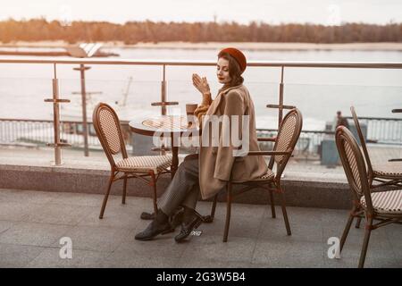 Gesamtplan einer nachdenklichen jungen Pariser Frau mit einem Kaffeebecher auf dem Tisch in roter Baskenmütze auf der Terrasse des Restaurants Stockfoto