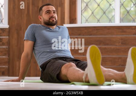 Entspannendes Sitzen lehnte sich zurück auf seine Arme junger glücklicher Fitness-Kerl macht Stretching-Übungen zu Hause. Amateur Sport Mann in worko Stockfoto