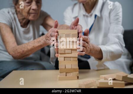 Jenga-Spiel. Thema sind Demenz, Altern und Spiele für alte Menschen. Kaukasische ältere Frau baut mit dem Hel einen Turm aus Holzblöcken Stockfoto