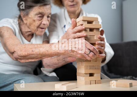 Jenga-Spiel. Thema sind Demenz, Altern und Spiele für alte Menschen. Kaukasische ältere Frau baut mit dem Hel einen Turm aus Holzblöcken Stockfoto