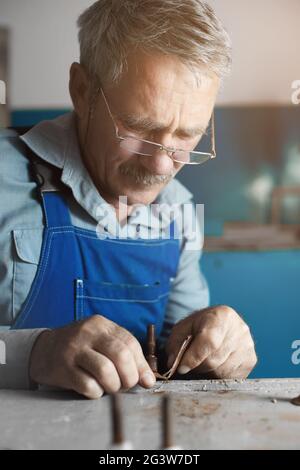 Ein älterer Meister mit Brille bei der Arbeit. Ein weißer alter Mann mit kaukasischem Aussehen sitzt an einem Tisch und arbeitet mit seinen Händen Stockfoto