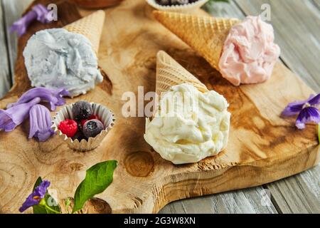 Vanille-, Heidelbeer- und Himbeereis in Waffelkegel auf Holzbrett mit Beeren und Blumen. Stockfoto
