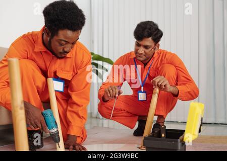 Ein Team von interracial Handwerkern in orangefarbenem Body hockend im Wohnzimmer und mit Handwerkzeugen beim Zusammenbauen des Tisches Stockfoto