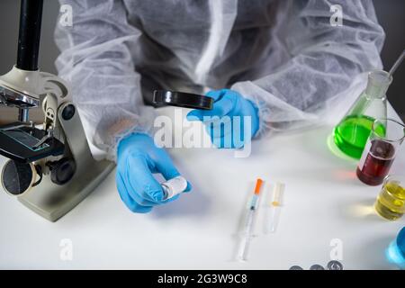 Wissenschaftler in Hazmat Anzug und Brille hält Impfstoffflasche und Lupe, suchen und forschen. Epidemiologie-Virustest. Stockfoto