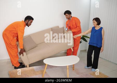 Ernsthafte reife asiatische Frau zeigt Platz für Sofa zu bewegen im Wohnzimmer, während in neues Haus bewegen Stockfoto
