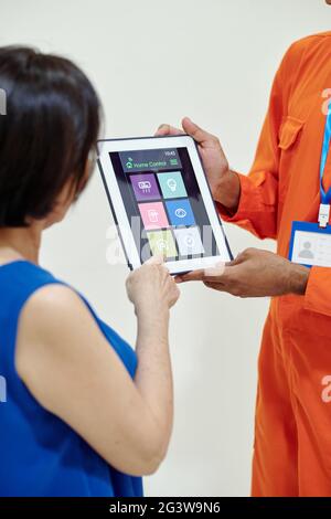 Techniker in orangefarbenem Anzug, der der Frau Symbole auf dem Tablet zeigt, während sie ihr beibringt, die App für die Heimsteuerung zu verwenden Stockfoto