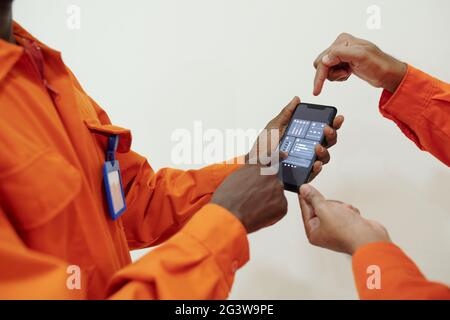 Nahaufnahme eines nicht erkennbaren schwarzen Mannes in orangefarbenem Anzug mithilfe der Smartphone-App mit einem Kollegen, um die Luftqualität aus der Ferne zu steuern Stockfoto
