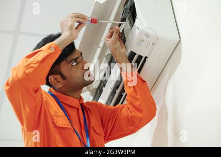 Konzentrierter junger indischer Ingenieur in Orange trägt beim Einrichten der Klimaanlage Schraubendreher Stockfoto