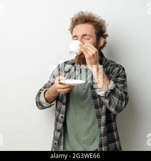 Gut aussehender Mann mit lockigem Haar und Bart trinkt Kaffee halten Tasse, trägt kariertes Langarm-Shirt isoliert auf weißem Rücken Stockfoto