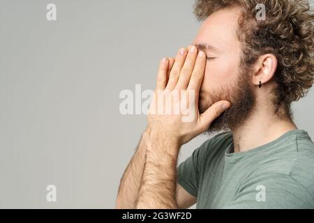 Hat Kopfschmerzen, junger Mann, der sein Gesicht berührt. Schöner junger Mann mit lockigen Haaren in olivem T-Shirt isoliert auf weißem Hintergrund Stockfoto