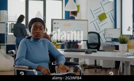 Junge schwarze Frau mit Behinderungen, ungültig, behindert Gehbehinderte, die die Kamera schauend verängstigt, traurig sitzend im Rollstuhl im Bürozimmer, beim Finanzprojekt mit dem Team arbeitend. Stockfoto