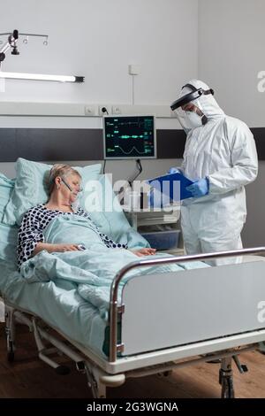 Ältere Patientin, die während des Coronavirus-Ausbruchs bewusstlos im Krankenhausbett lag und mit Hilfe der Sauerstoffmaske atmete. Arzt mit psa-Anzug, um Infektionen zu verhindern. Stockfoto