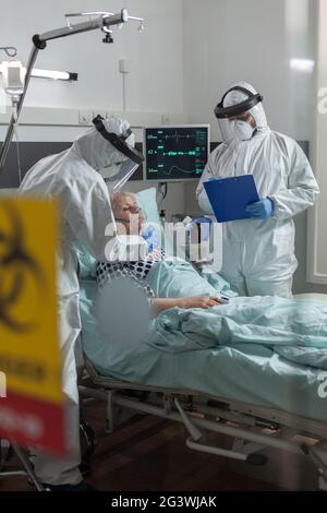 Medizinische Krankenschwester in PSA-Anzug, die im Zuge einer globalen Pandemie mit Coronavirus eine Sauerstoffmaske an einen älteren Patienten legt, und der Arzt notiert sich in der Zwischenablage. Stockfoto