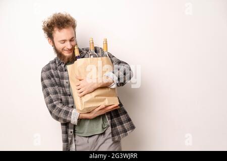 Ein junger Mann, der eine Einkaufstasche aus Papier mit lachenden Weinflaschen trägt und darauf herabblickt. Hübscher junger Mann mit lockigen Haaren in Oli Stockfoto