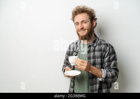 Lächelnder, gutaussehender Mann mit lockigem Haar und Bart, trinkt Kaffee und trägt ein kariertes Langarm-Hemd, isoliert auf weißem Ba Stockfoto