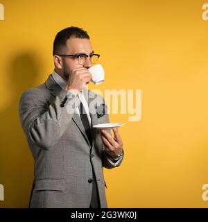 Hübscher junger bärtiger Geschäftsmann in einer Brille in formeller Kleidung mit einer Tasse Kaffee, der seitlich isoliert auf gelbem B steht Stockfoto