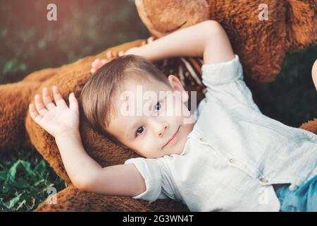 Ein schöner kleiner Junge liegt mit seinem Lieblings-Plüschtier auf dem Gras im Park und schaut auf die Kamera. Stockfoto