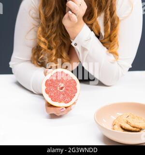 Eine dicke junge Frau, die eine frische Grapefruit in der Hand hält und zu Hause mit langen, lockigen, blonden Haaren auf einem Küchentisch steht. Dietin Stockfoto