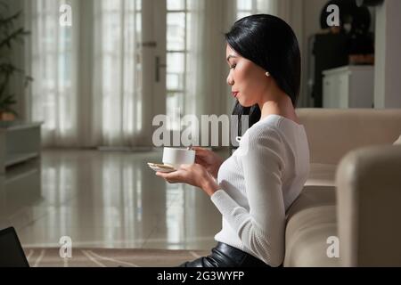 Ruhige, elegante junge asiatische Frau mit schwarzen Haaren, die auf dem Boden sitzen und sich auf dem Sofa lehnen, während sie im Wohnzimmer Kaffee trinkt Stockfoto
