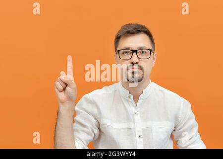 Junger bärtiger, nachdenklicher Mann in einer Brille, der mit dem Finger nach oben zeigt, zeigt an, dass er den Raum kopiert, oranger Studiohintergrund Stockfoto