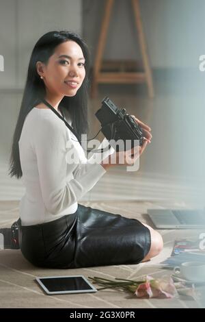 Porträt eines glücklichen jungen vietnamesischen Fotografen in Schwarz-Weiß-Kleidung, die auf dem Teppich sitzt und eine Vintage-Kamera hält Stockfoto