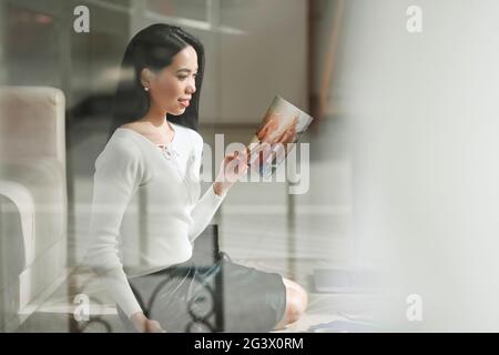 Glückliche junge asiatische Frau in weißem Hemd sitzt auf dem Boden und Blick auf Foto, während Erinnerungen zu genießen Stockfoto
