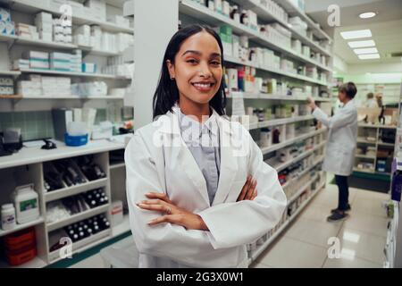 Porträt eines jungen afroamerikanischen Apothekers, der in einem Chemiker zwischen den Gängen steht, mit einem Kollegen, der im Hintergrund arbeitet Stockfoto
