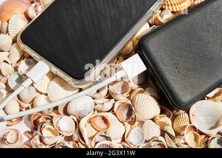 Powerbank lädt ein Smartphone auf Muscheln. Nahaufnahme, Draufsicht. Stockfoto