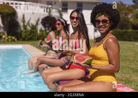 Eine vielfältige Gruppe von Freundinnen, die Wassermelone essen und am Pool sitzen und sich unterhalten Stockfoto