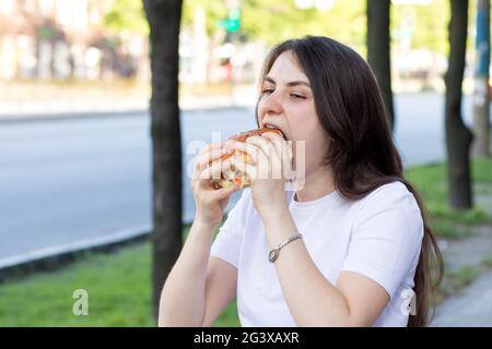 Brünette Frau überfrisst auf einem Burger auf der Straße. Völlerei, überschüssige Kalorien und Bulimie. Stockfoto