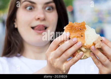 Brünette Frau überfrisst auf einem Burger auf der Straße. Völlerei, überschüssige Kalorien und Bulimie. Stockfoto