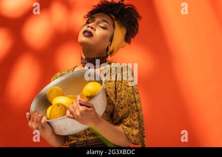 afroamerikanische Frau in Bluse und Kopftuch mit Metallschale mit Zitronen auf Orange Stockfoto