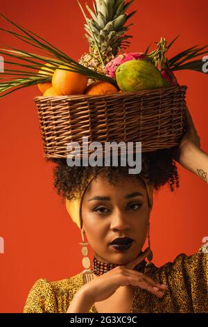 Porträt einer ernsthaften jungen afroamerikanischen Frau, die einen Korb mit exotischen Früchten auf dem Kopf auf Rot hält Stockfoto