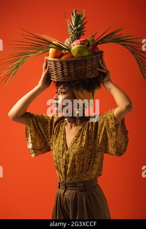 Ernsthafte junge afroamerikanische Frau, die Korb mit exotischen Früchten auf dem Kopf auf Rot hält Stockfoto