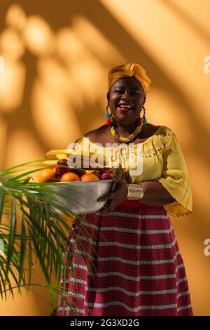 afroamerikanische Frau mittleren Alters in gelber Bluse und Turban mit Metallschüssel mit Früchten auf Orange Stockfoto