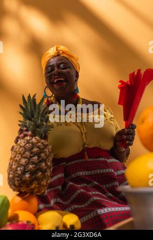 Aus der unteren Perspektive sehen Sie eine afroamerikanische Frau mittleren Alters, die Früchte mit Ananas verkauft und einen Fächer in den Händen auf Orange hält Stockfoto