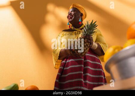 Aus der Perspektive einer lächelnden afroamerikanischen Frau mittleren Alters, die Früchte mit Ananas in der Hand auf Orange verkauft Stockfoto