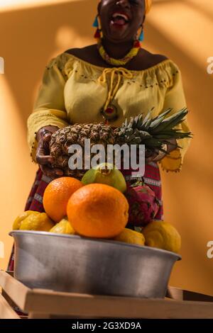 Aus der unteren Perspektive eine fröhliche afroamerikanische Frau mittleren Alters, die Früchte mit Ananas in den Händen auf Orange verkauft Stockfoto