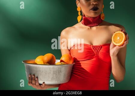 Teilansicht einer afroamerikanischen Frau in rotem Badeanzug mit geschnittener Orange und Metallschale mit Früchten auf Grün Stockfoto